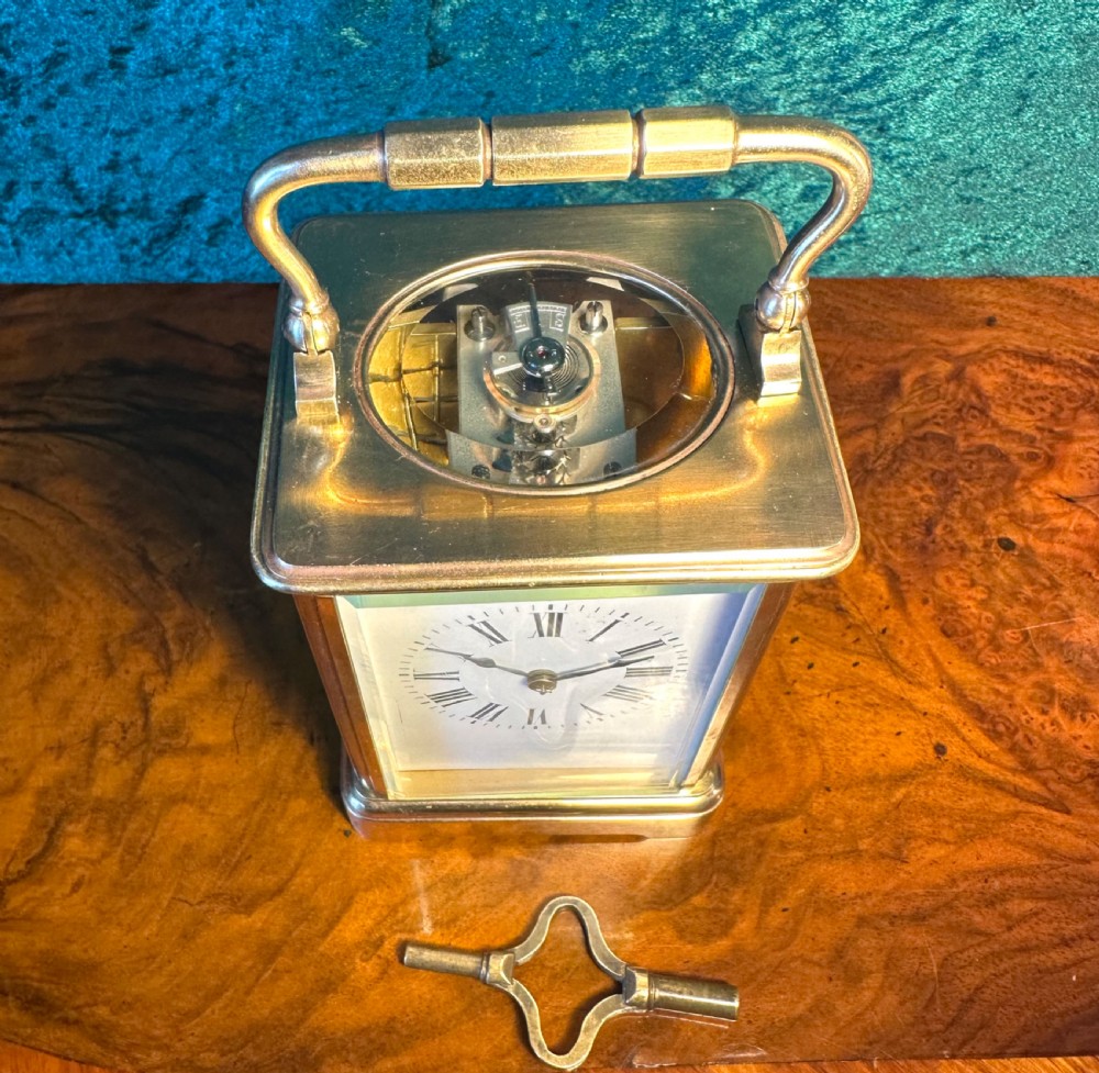 A rectangular brass carriage clock featuring glass side panels, a white enamel dial with black Roman numerals, and a top carrying handle. View VI
