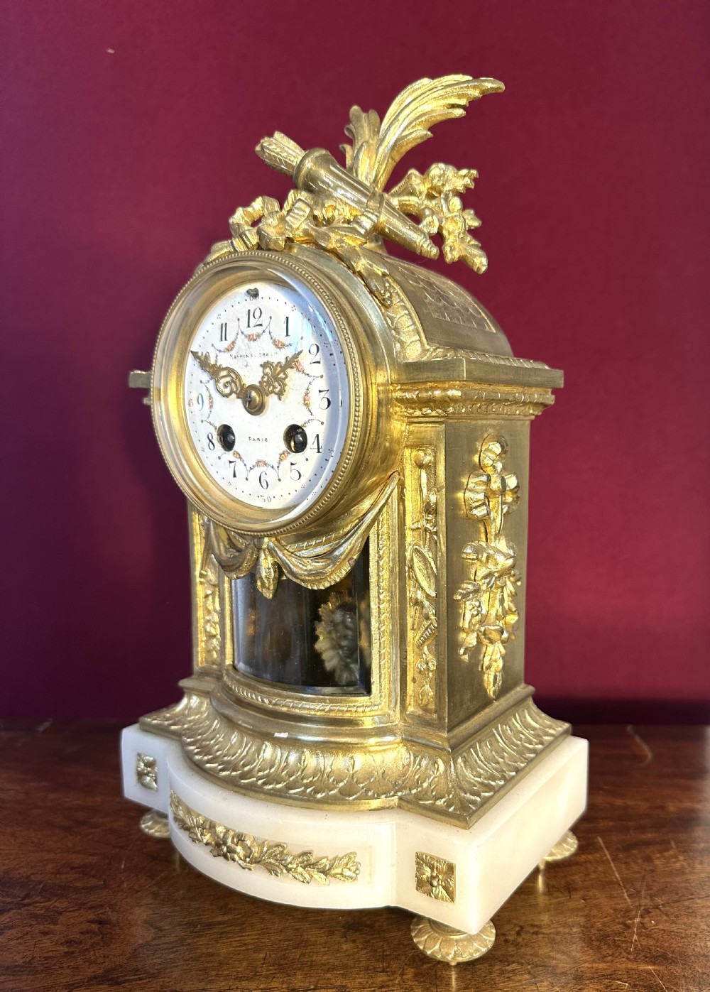 An antique late 19th-century French gilt bronze and white alabaster mantel clock featuring a hand-painted enamel dial, floral swags, and a quiver motif on top.