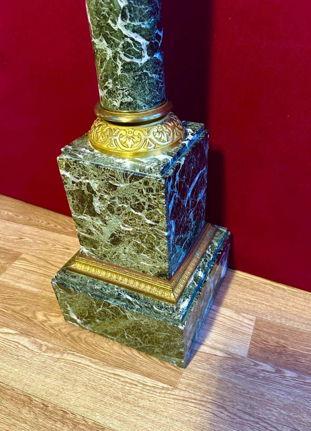 A tall dark green veined marble column pedestal featuring a square top, gilt bronze Corinthian capital, cylindrical shaft, and a stepped square base. View V