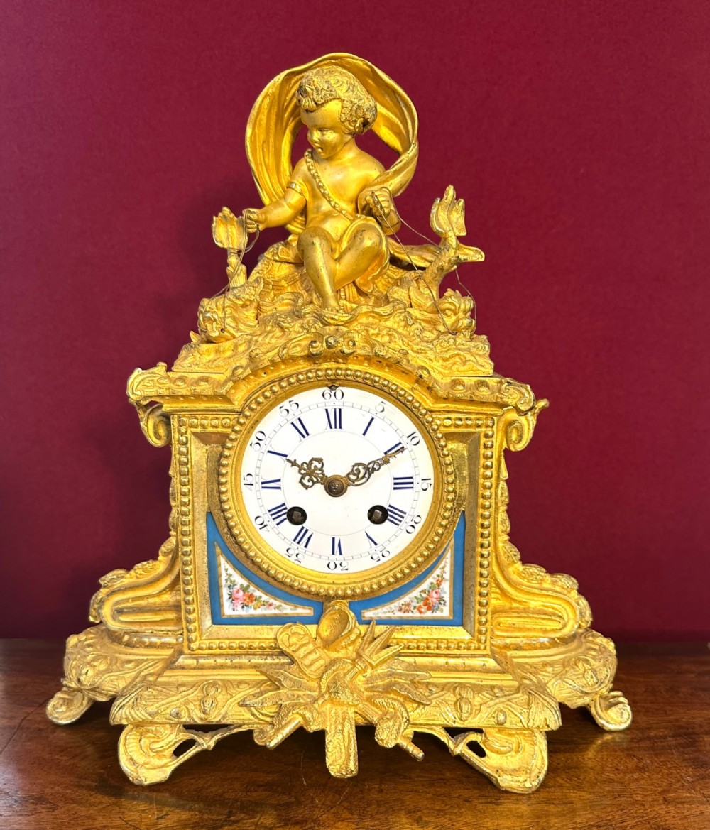 A late 19th-century French gilt bronze mantel clock crowned with a seated cherub figure and featuring an enamel dial with blue porcelain panels.