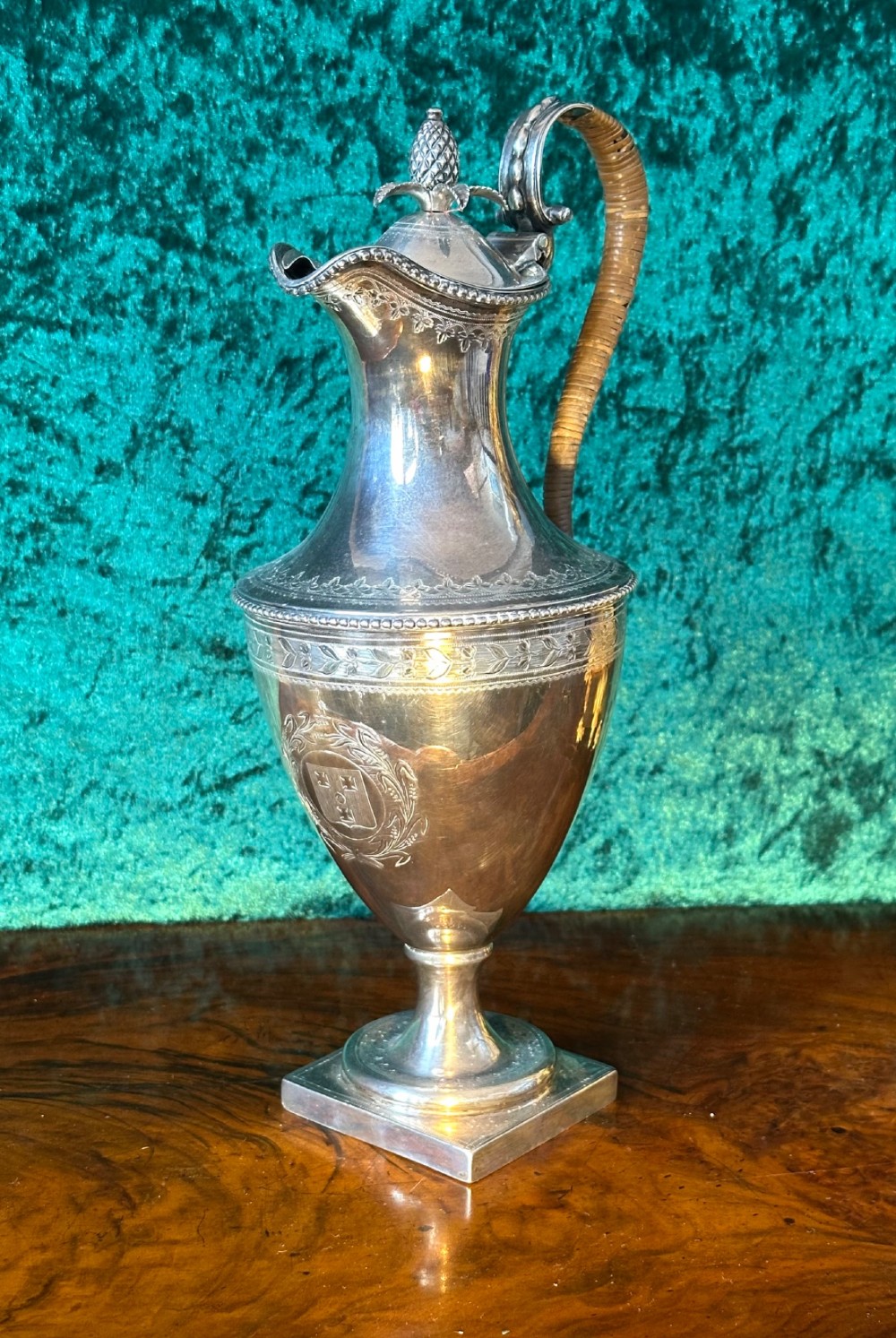 A Georgian silver ewer with an urn-shaped body, engraved heraldic coat of arms, pineapple finial, and a cane-wrapped handle resting on a square base.