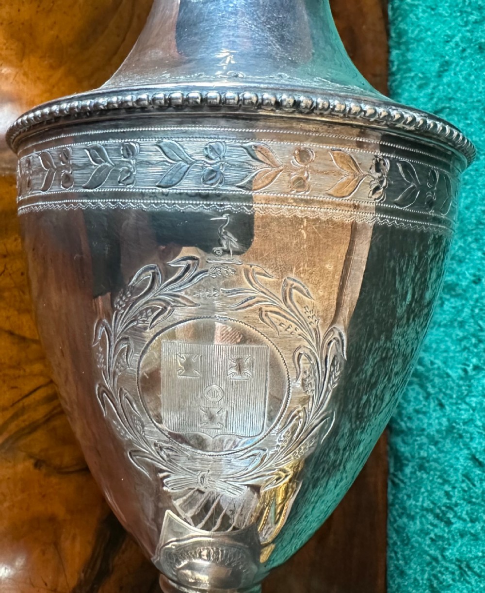 A Georgian silver ewer with an urn-shaped body, engraved heraldic coat of arms, pineapple finial, and a cane-wrapped handle resting on a square base. View IV
