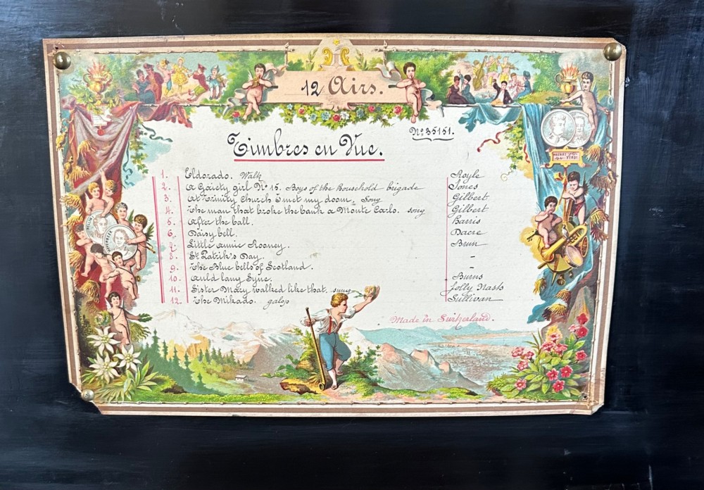 An open Victorian cylinder music box housed in a dark veneered wooden case featuring six exposed bells and an illustrated tune sheet inside the lid. View VII