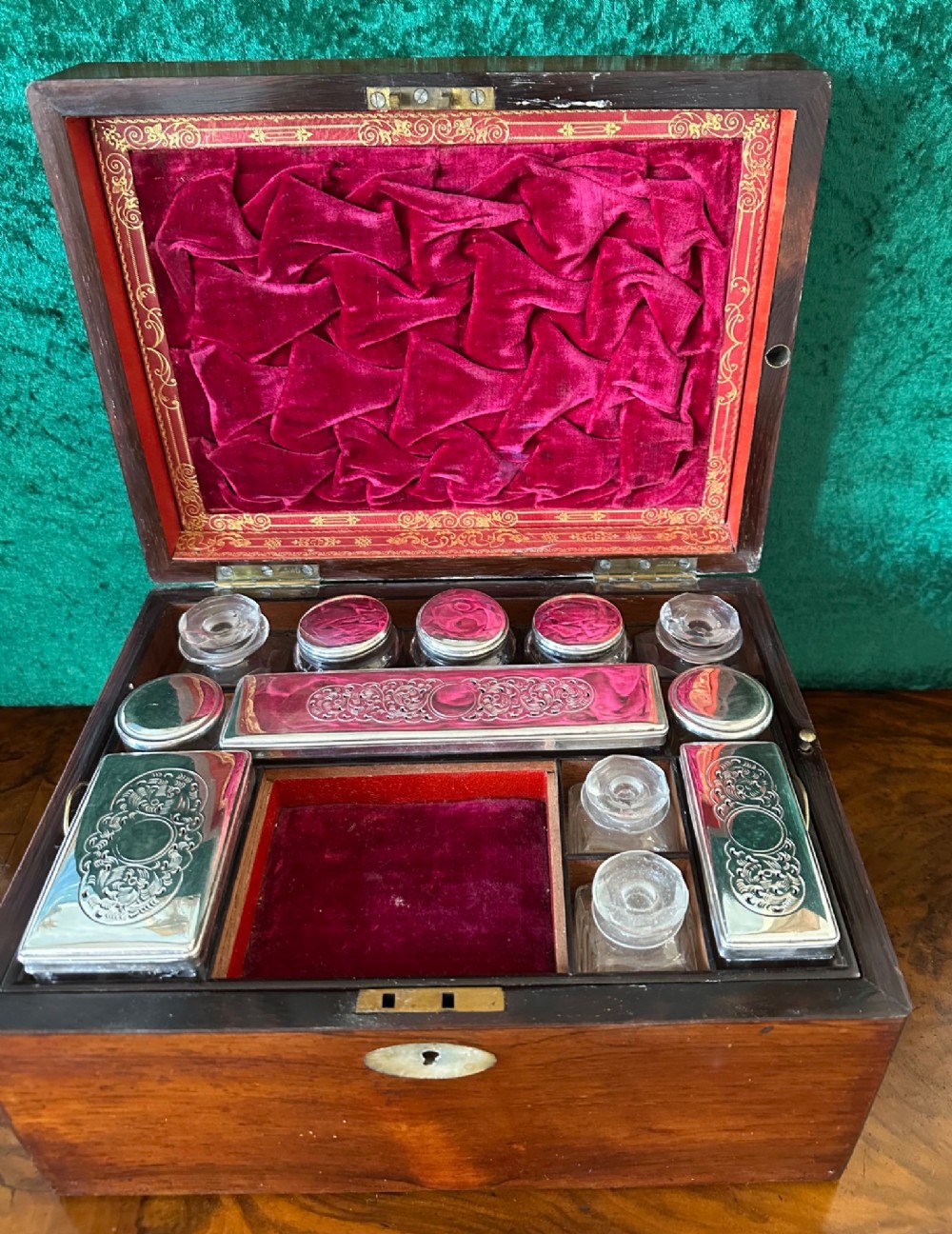 An open Victorian wooden travelling dressing case featuring vibrant red velvet lining, glass toiletry jars, engraved silver lids, and small compartments.