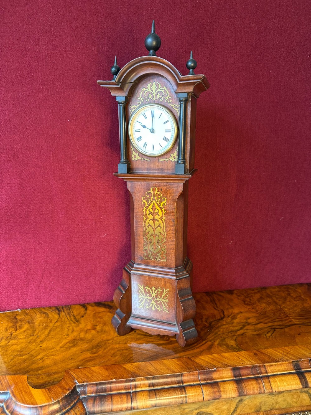 A wooden miniature grandfather clock featuring brass foliate inlay, an arched hood with three turned finials, black side columns, and a cream dial face with Roman numerals. View II
