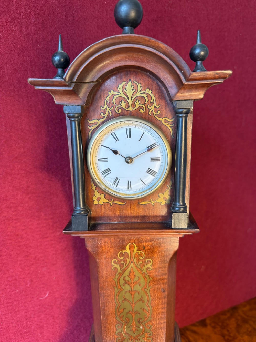 A wooden miniature grandfather clock featuring brass foliate inlay, an arched hood with three turned finials, black side columns, and a cream dial face with Roman numerals. View III