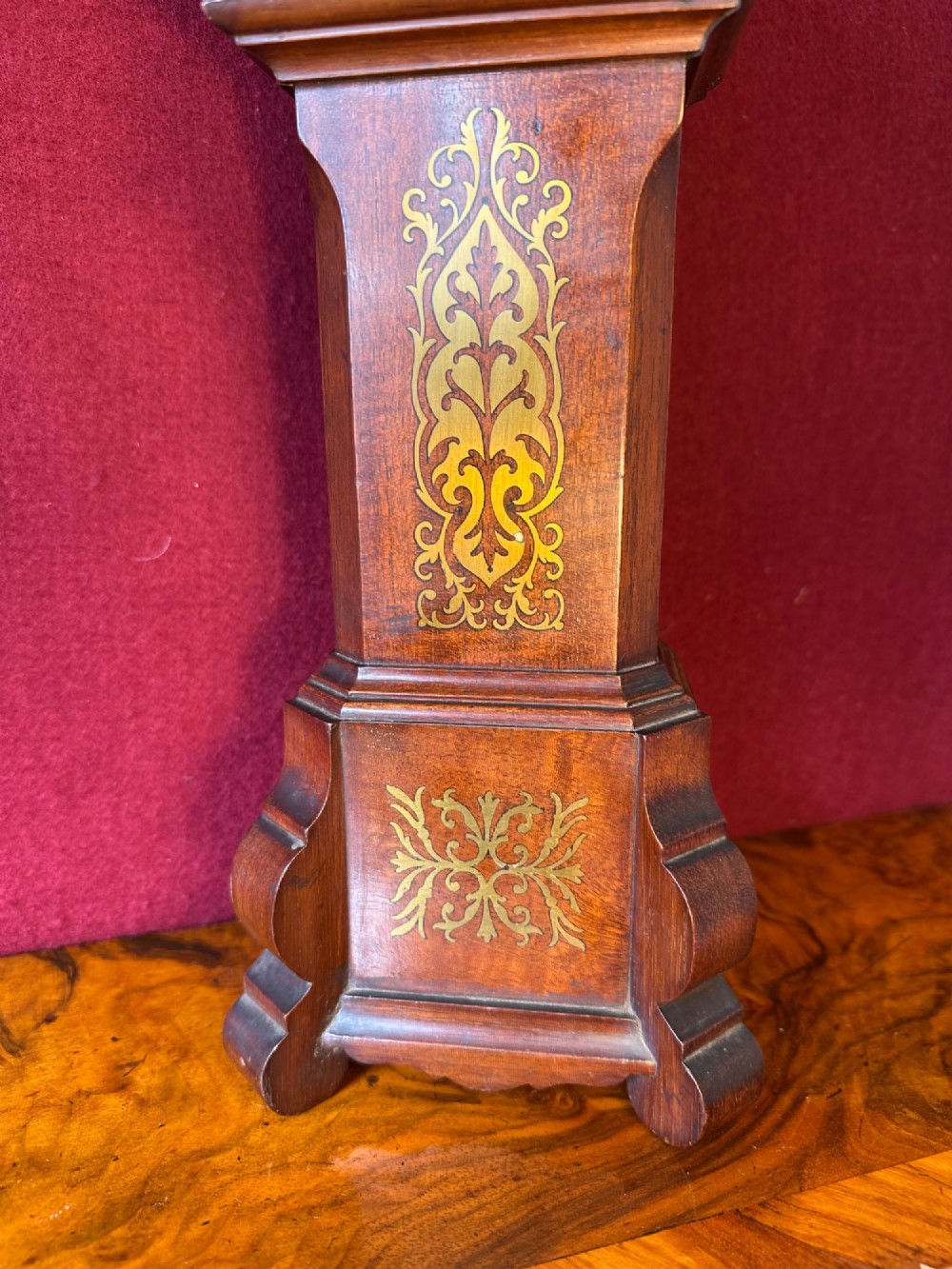 A wooden miniature grandfather clock featuring brass foliate inlay, an arched hood with three turned finials, black side columns, and a cream dial face with Roman numerals. View IV