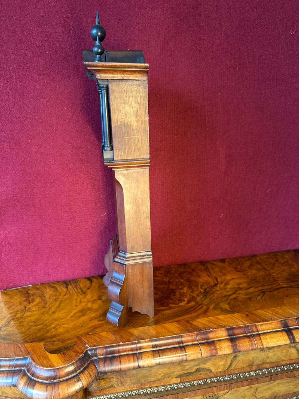 A wooden miniature grandfather clock featuring brass foliate inlay, an arched hood with three turned finials, black side columns, and a cream dial face with Roman numerals. View V