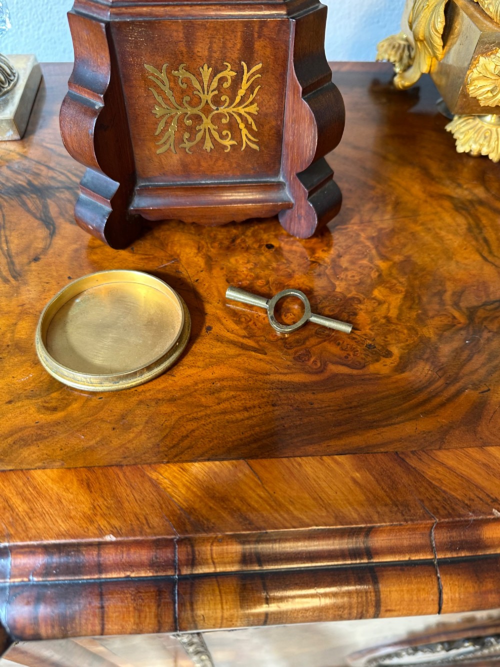 A wooden miniature grandfather clock featuring brass foliate inlay, an arched hood with three turned finials, black side columns, and a cream dial face with Roman numerals. View VII