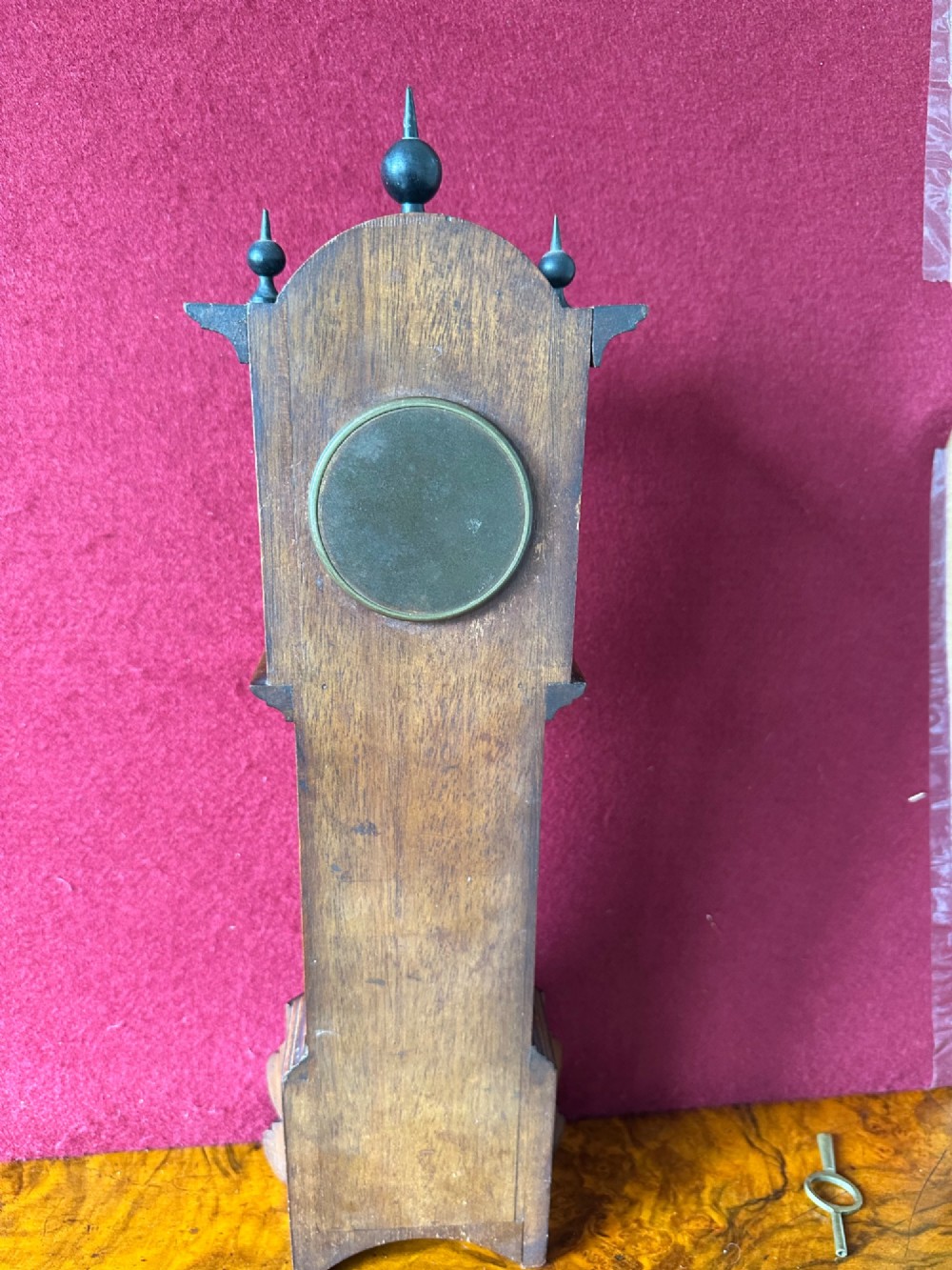 A wooden miniature grandfather clock featuring brass foliate inlay, an arched hood with three turned finials, black side columns, and a cream dial face with Roman numerals. View IX