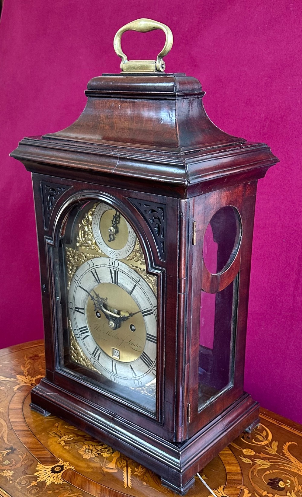A mid-18th-century Georgian mahogany bracket clock featuring an upper brass carrying handle, an arched glass front door, and an engraved brass dial with a silvered chapter ring. View II