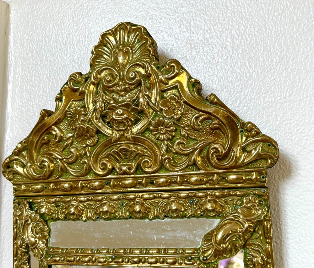An antique repousse brass marginal mirror featuring an ornate floral pediment crest and bevelled glass borders. View II