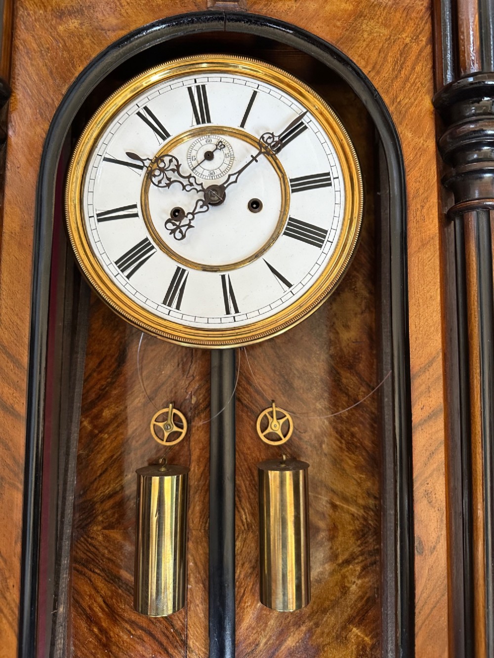 Late 19th-century Victorian walnut wall clock with ebonised columns, top cresting with a carved head motif, two brass weights, pendulum, and a white enamelled Roman numeral dial. View II