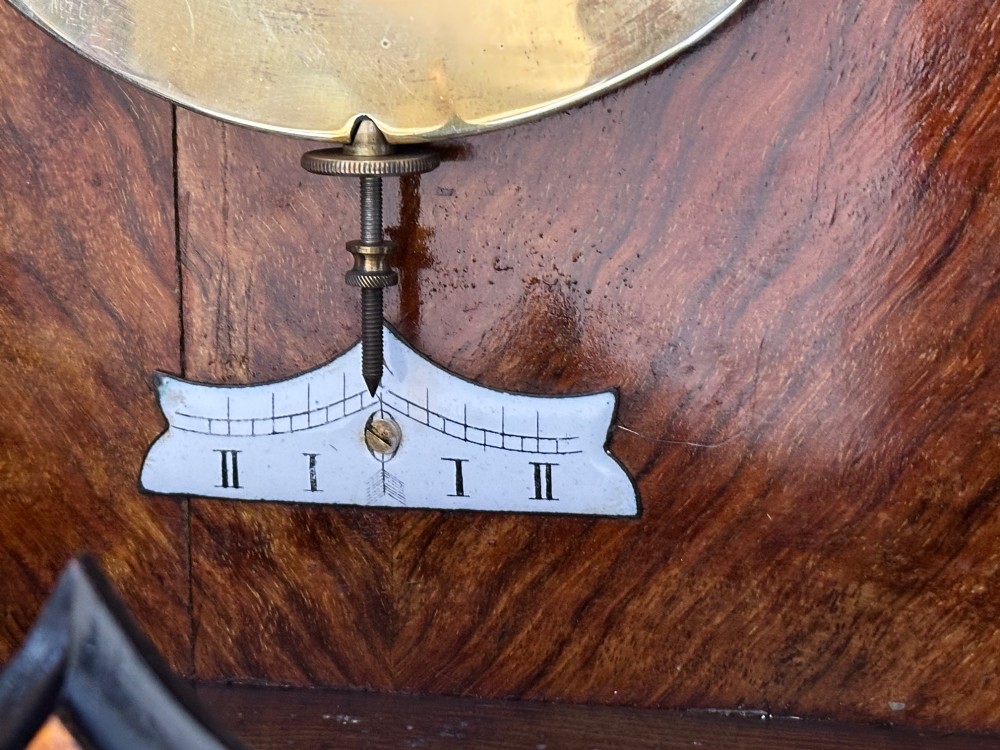 Late 19th-century Victorian walnut wall clock with ebonised columns, top cresting with a carved head motif, two brass weights, pendulum, and a white enamelled Roman numeral dial. View V