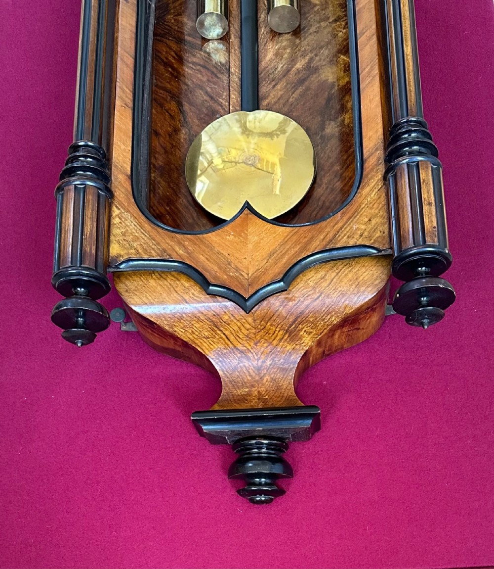 Late 19th-century Victorian walnut wall clock with ebonised columns, top cresting with a carved head motif, two brass weights, pendulum, and a white enamelled Roman numeral dial. View VII