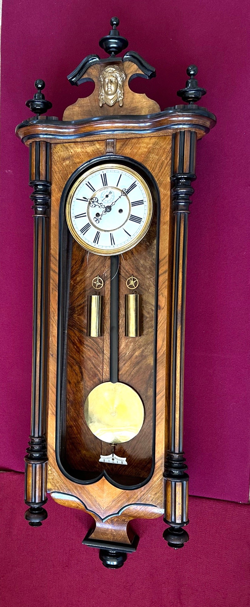 Late 19th-century Victorian walnut wall clock with ebonised columns, top cresting with a carved head motif, two brass weights, pendulum, and a white enamelled Roman numeral dial. View XI
