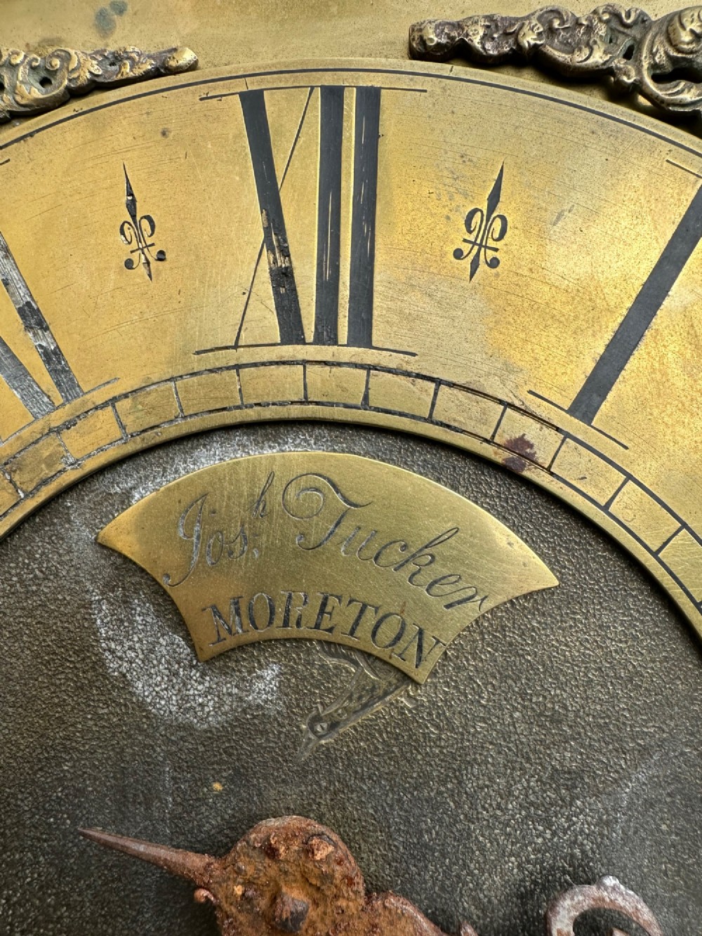 A square eighteenth-century brass clock dial with cast corner spandrels, Roman numerals, engraved centre detailing, and rusty iron chains. View II