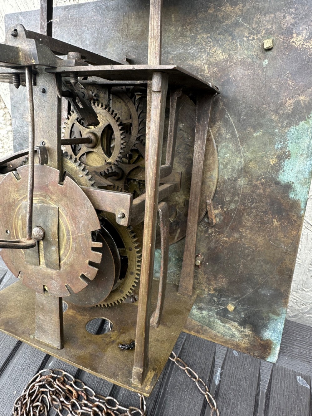 A square eighteenth-century brass clock dial with cast corner spandrels, Roman numerals, engraved centre detailing, and rusty iron chains. View VI