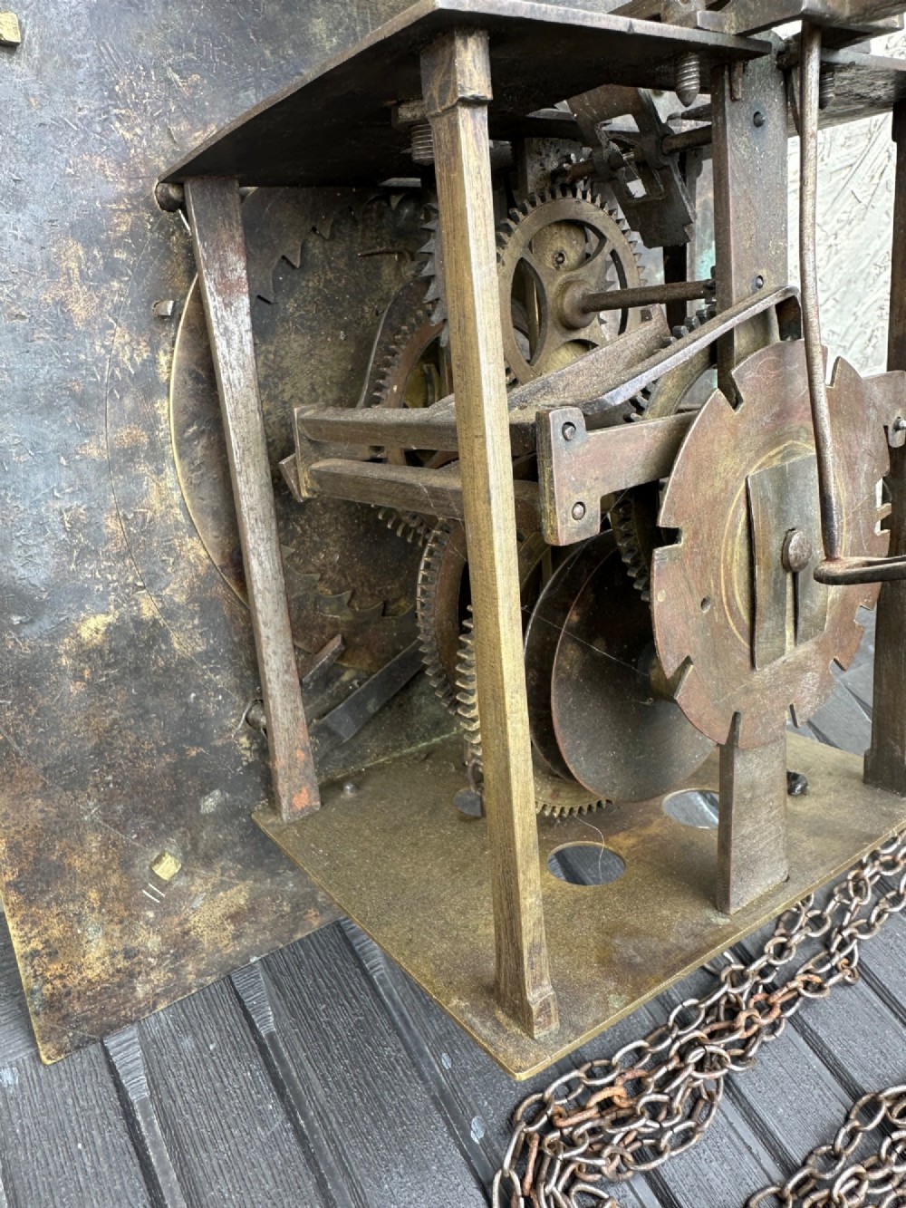 A square eighteenth-century brass clock dial with cast corner spandrels, Roman numerals, engraved centre detailing, and rusty iron chains. View VII