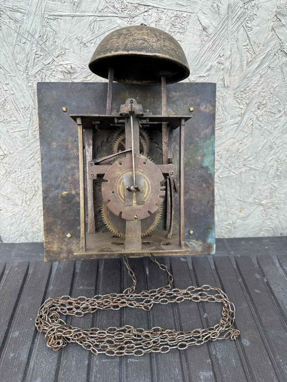 A square eighteenth-century brass clock dial with cast corner spandrels, Roman numerals, engraved centre detailing, and rusty iron chains. View VIII