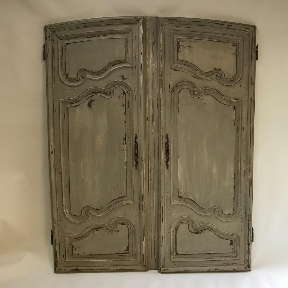 A pair of grey painted wooden doors featuring carved Louis XV style moulded panels and a distressed paint finish with central iron escutcheons.