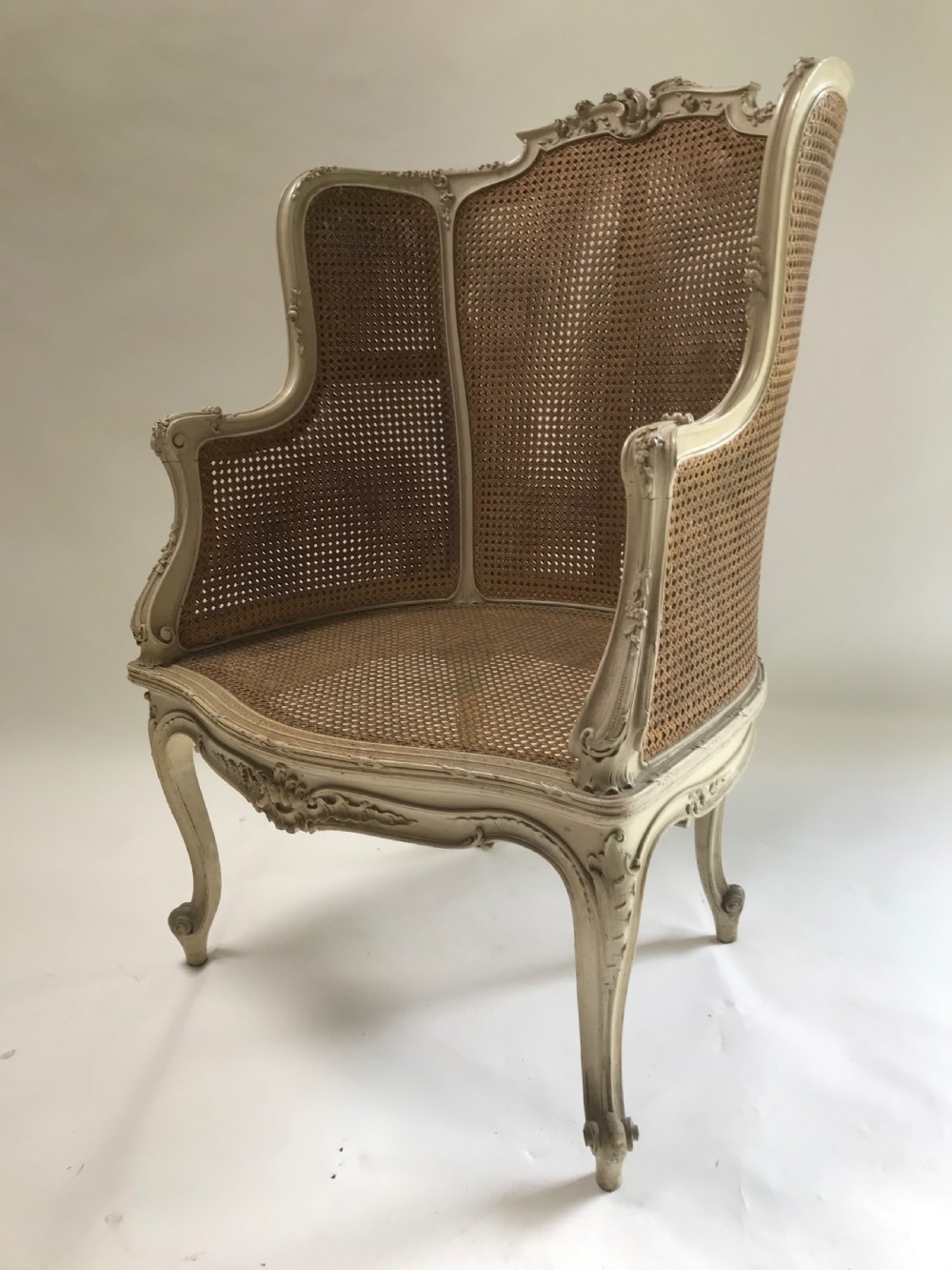 A French Louis XV style painted armchair featuring hand-woven cane back and sides, carved floral motifs, cream distressed paint finish, and cabriole legs.