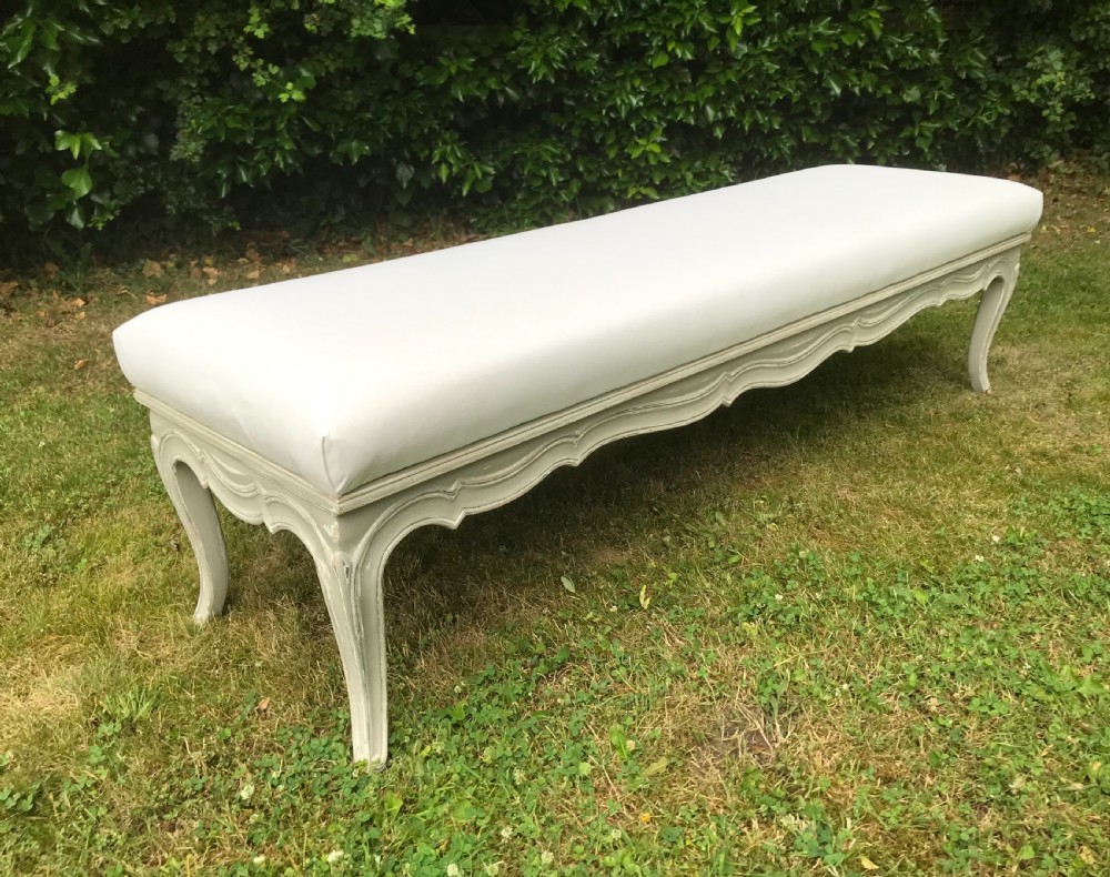 A long French style upholstered stool featuring a painted grey carved wooden frame with cabriole legs and a plain cream fabric top.