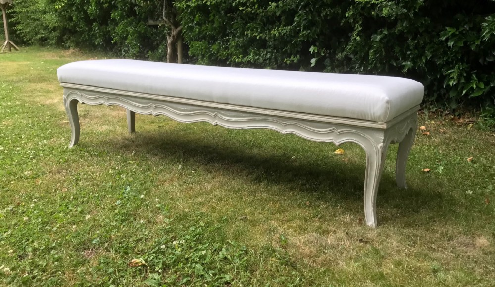 A long French style upholstered stool featuring a painted grey carved wooden frame with cabriole legs and a plain cream fabric top. View II