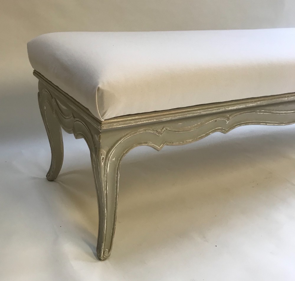 A long French style upholstered stool featuring a painted grey carved wooden frame with cabriole legs and a plain cream fabric top. View IV