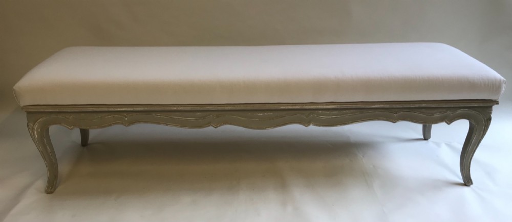 A long French style upholstered stool featuring a painted grey carved wooden frame with cabriole legs and a plain cream fabric top. View V