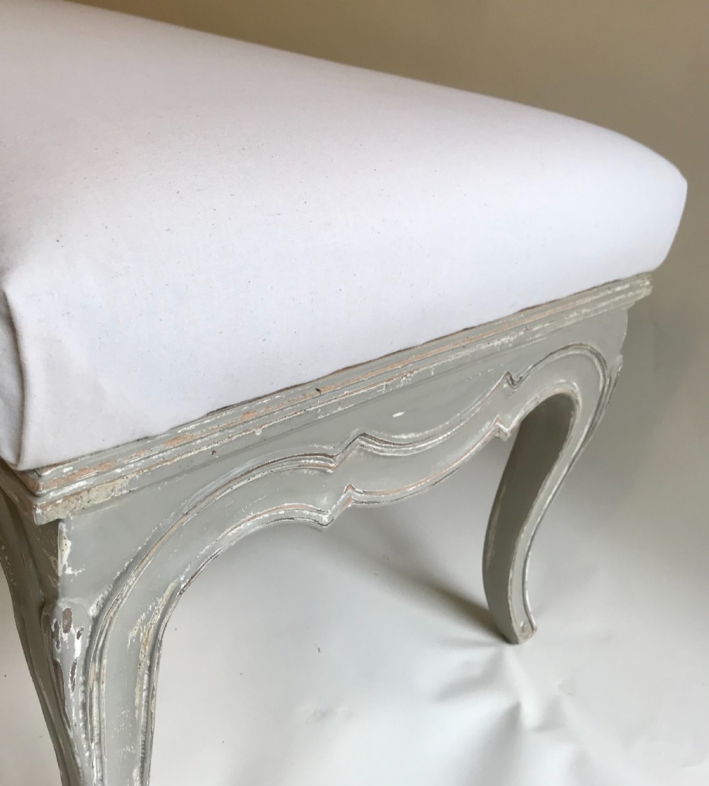 A long French style upholstered stool featuring a painted grey carved wooden frame with cabriole legs and a plain cream fabric top. View VI