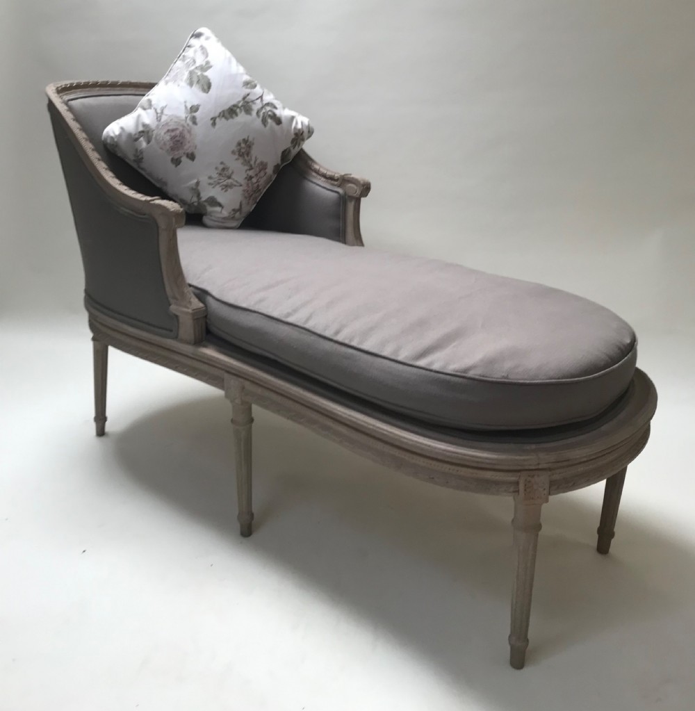 A French Louis XVI style upholstered daybed featuring a carved taupe-grey timber frame, fluted legs, a grey cushion seat, and a floral accent pillow.