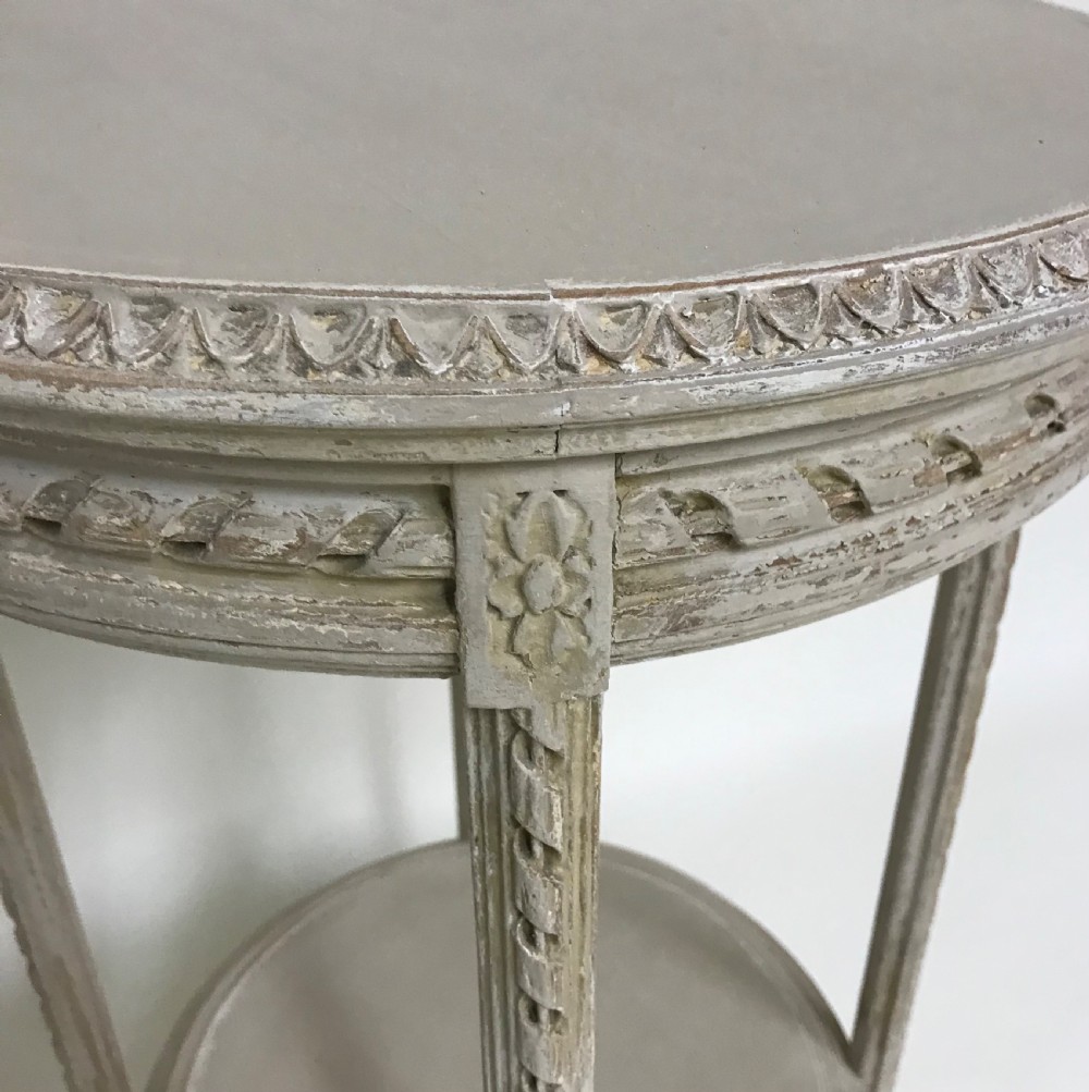 A circular grey painted wooden side table featuring a carved frieze, fluted legs, and a lower tier. View II