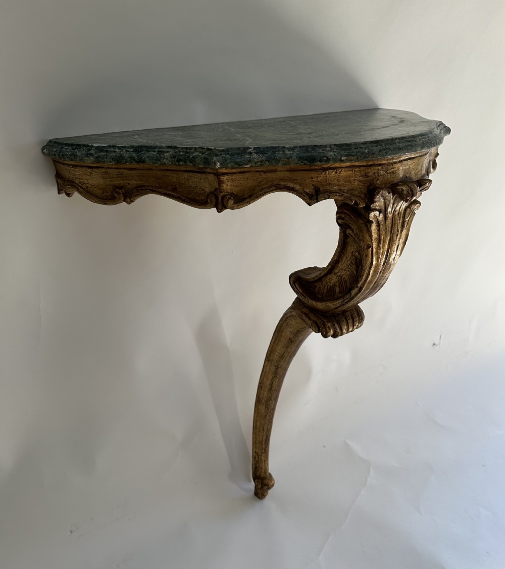 A wall-mounted Italian gilded console table featuring a single curved carved leg and a dark green marble top.
