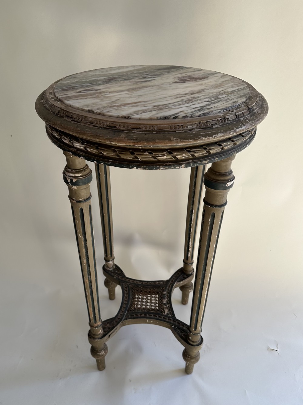 A circular French Louis XVI style occasional table featuring a veined marble top, distressed grey paint with gilt details, fluted legs, and a low X-frame stretcher.