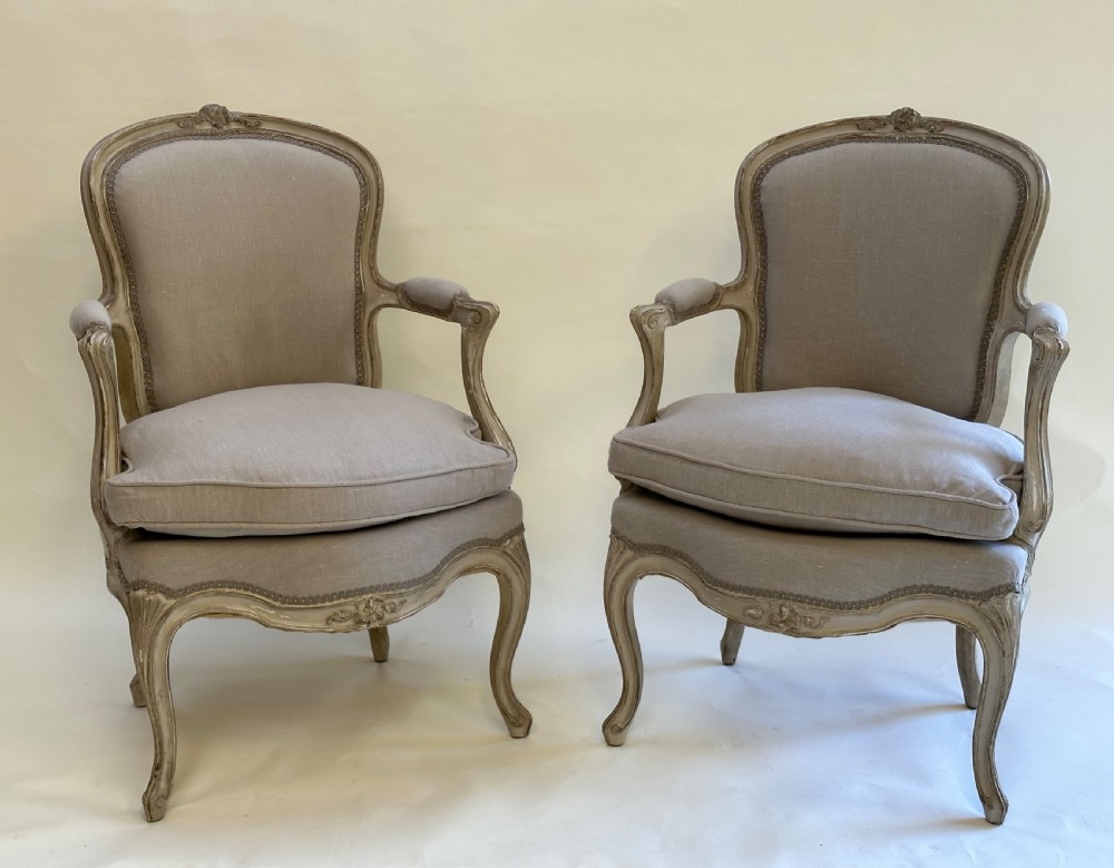 A pair of French Louis XV style open armchairs with grey painted carved wooden frames and neutral linen upholstery.