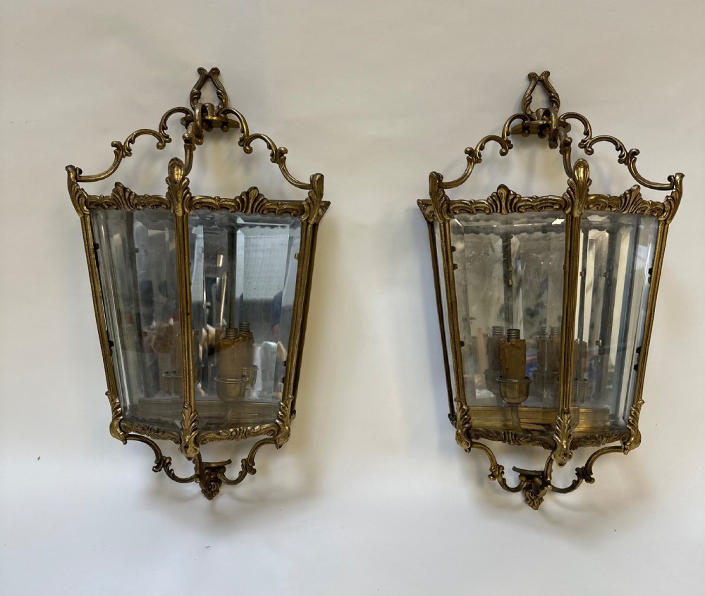 A pair of French antique gilt brass and bevelled glass wall lanterns featuring ornamental scrollwork crests and internal candle-style light fittings.