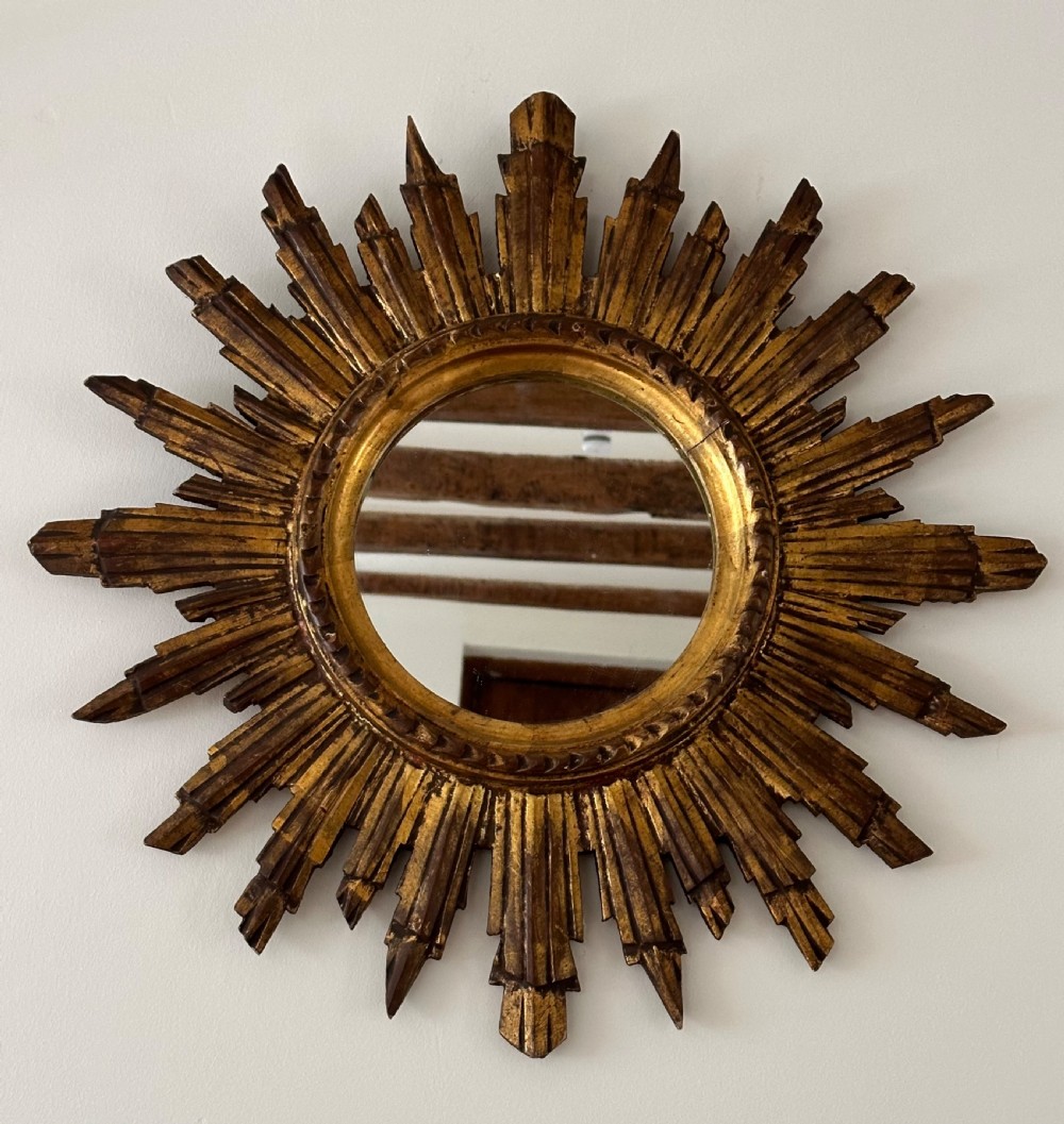 A circular wall mirror set within a gilded wooden frame featuring radiating, multi-tiered sunburst rays.