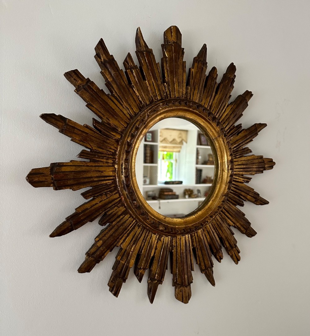 A circular wall mirror set within a gilded wooden frame featuring radiating, multi-tiered sunburst rays. View III