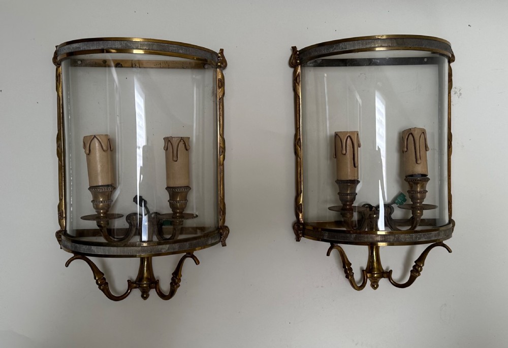 A pair of French brass wall lanterns featuring curved glass fronts, scrollwork lower supports, and twin candle sleeves within each frame.