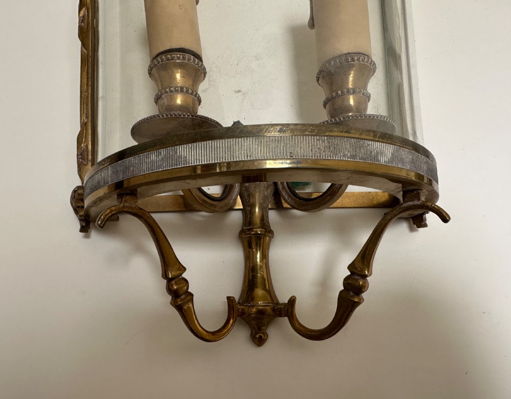 A pair of French brass wall lanterns featuring curved glass fronts, scrollwork lower supports, and twin candle sleeves within each frame. View V
