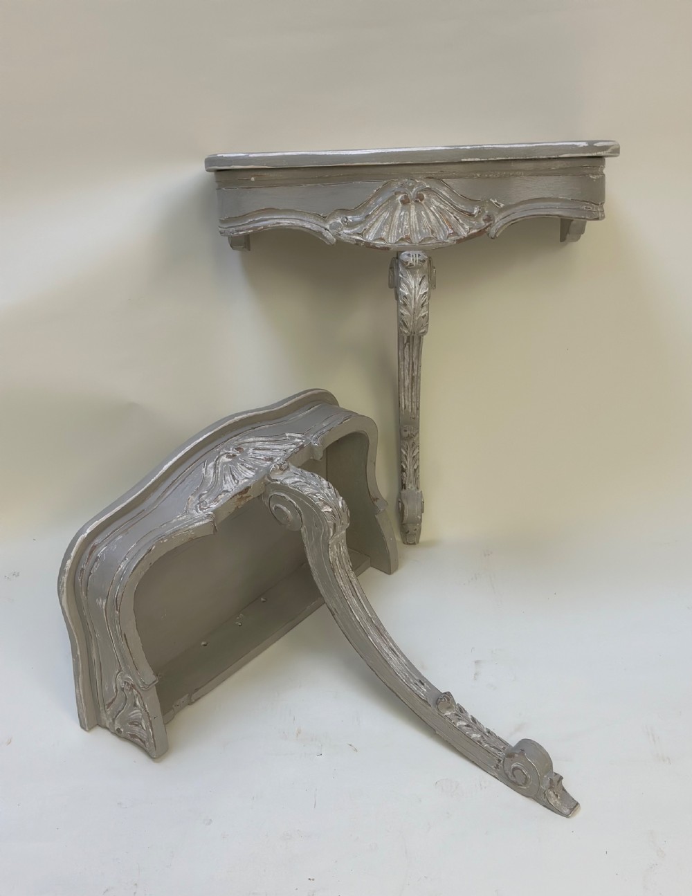 A pair of distressed grey painted French wall-mounted console tables featuring carved scallop shell motifs and single scrolled legs.