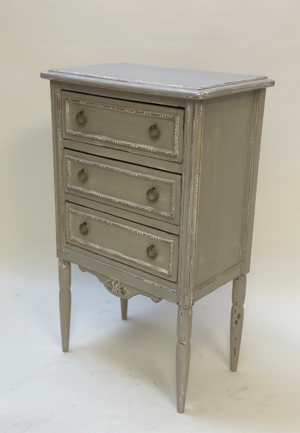 A small grey painted French-style chest with three drawers, brass ring handles, distressed edges, and tall turned legs.