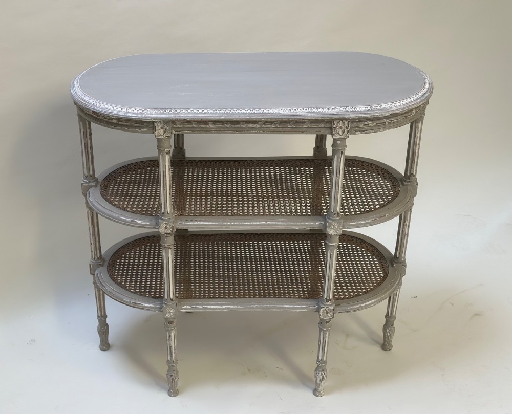 An oval French-style tiered side table painted in distressed grey with fluted legs, carved rosettes, and two cane lower shelves.