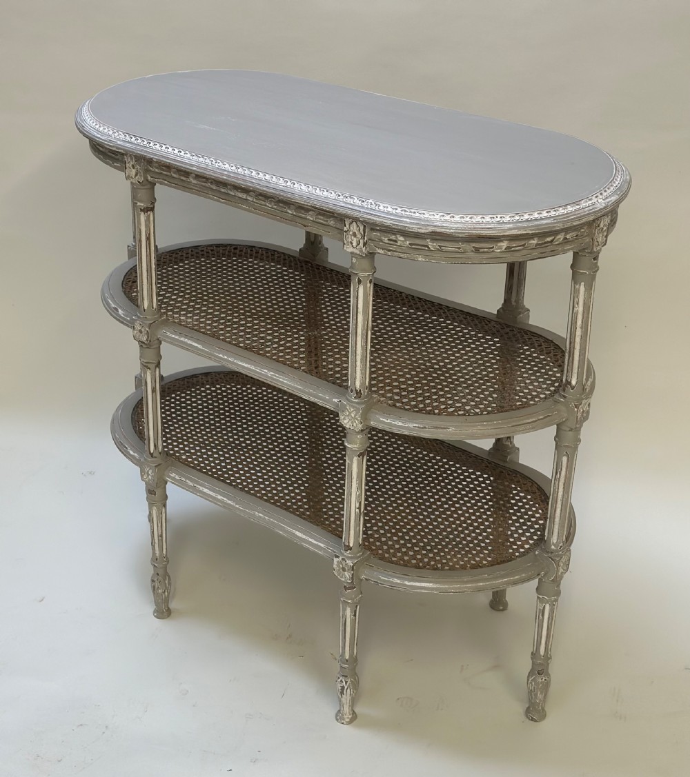 An oval French-style tiered side table painted in distressed grey with fluted legs, carved rosettes, and two cane lower shelves. View V