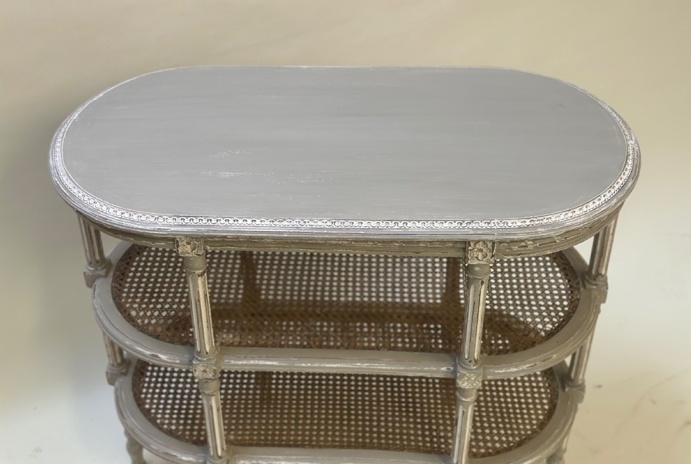 An oval French-style tiered side table painted in distressed grey with fluted legs, carved rosettes, and two cane lower shelves. View VIII