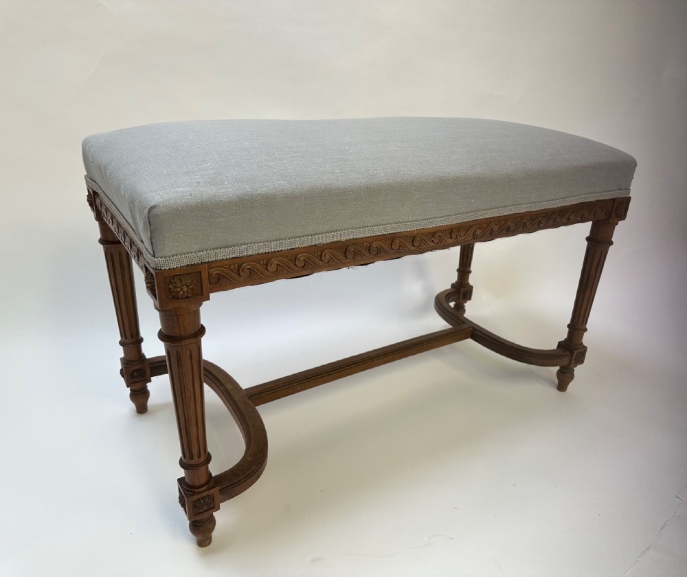 A rectangular French Neoclassical style stool featuring light grey linen upholstery, carved frieze woodwork, fluted legs, and a curved stretcher.