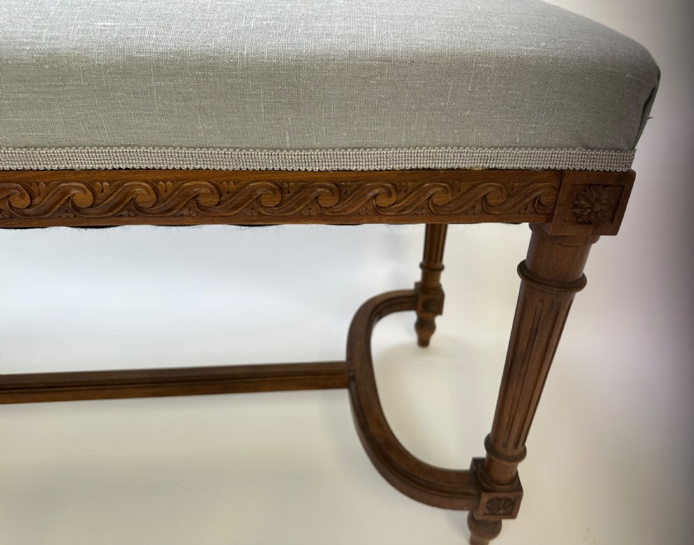 A rectangular French Neoclassical style stool featuring light grey linen upholstery, carved frieze woodwork, fluted legs, and a curved stretcher. View IV