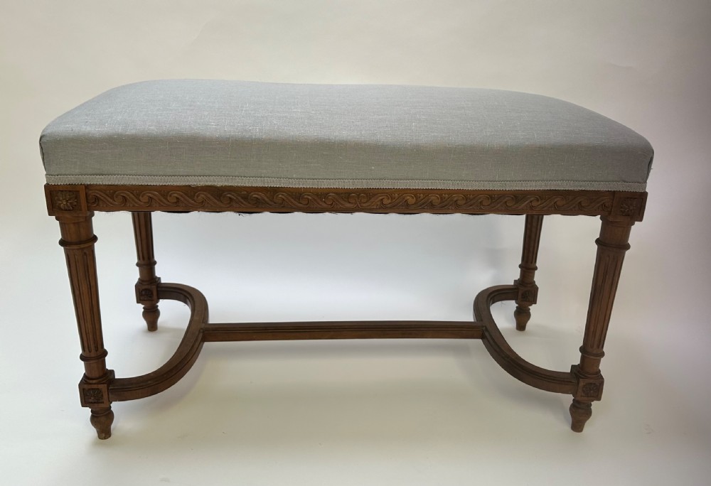 A rectangular French Neoclassical style stool featuring light grey linen upholstery, carved frieze woodwork, fluted legs, and a curved stretcher. View VI