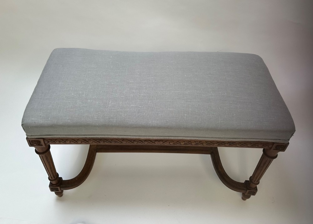 A rectangular French Neoclassical style stool featuring light grey linen upholstery, carved frieze woodwork, fluted legs, and a curved stretcher. View IX